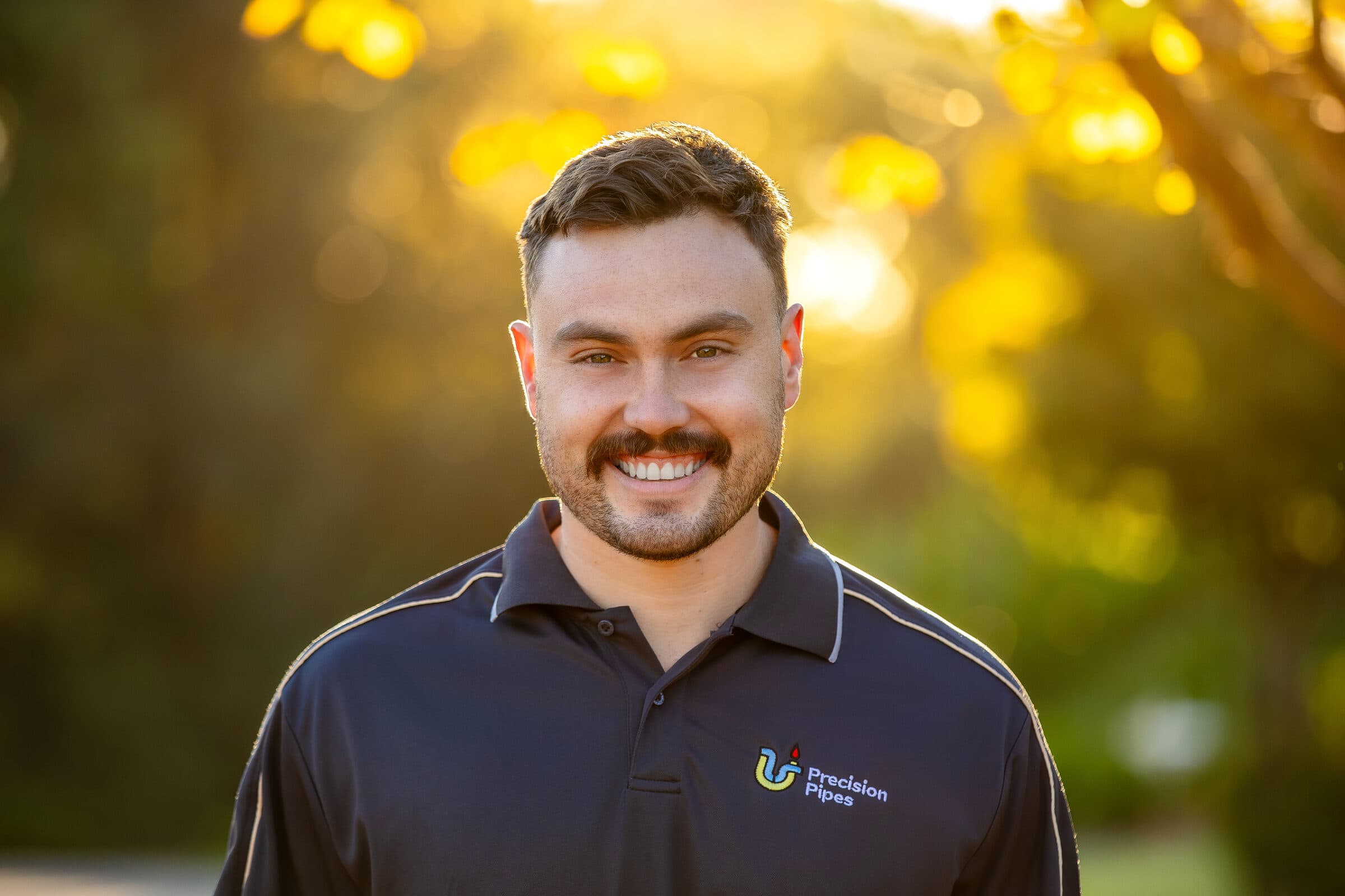 Colin, the owner of Precision Pipes, in a branded navy polo, photographed on a tree-lined street at golden hour