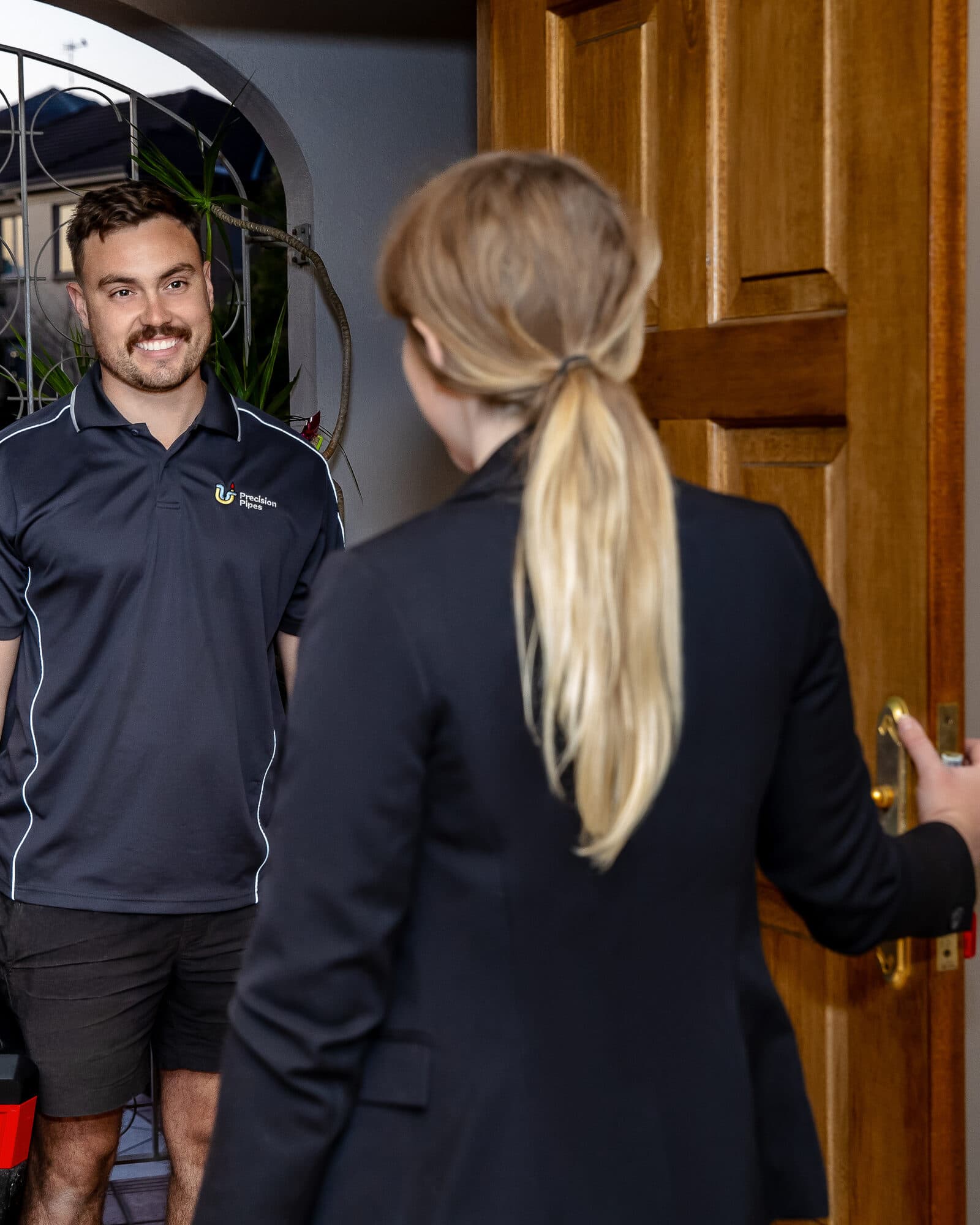 A Precision Pipes plumber arriving at the front door of a home with his toolbox, greeted by the customer