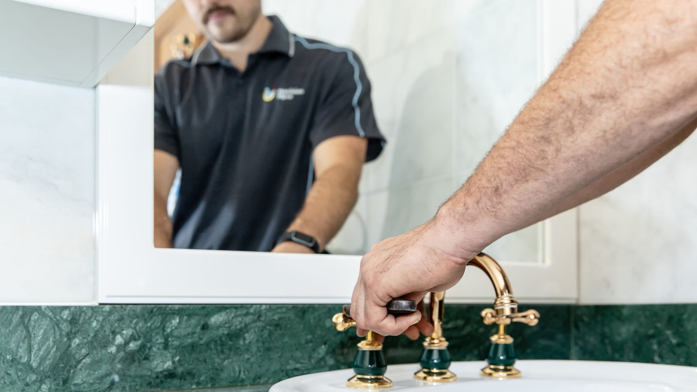 Close-up of a Precision Pipes plumber’s hand turning a gold basin tap, reflected in the mirror behind